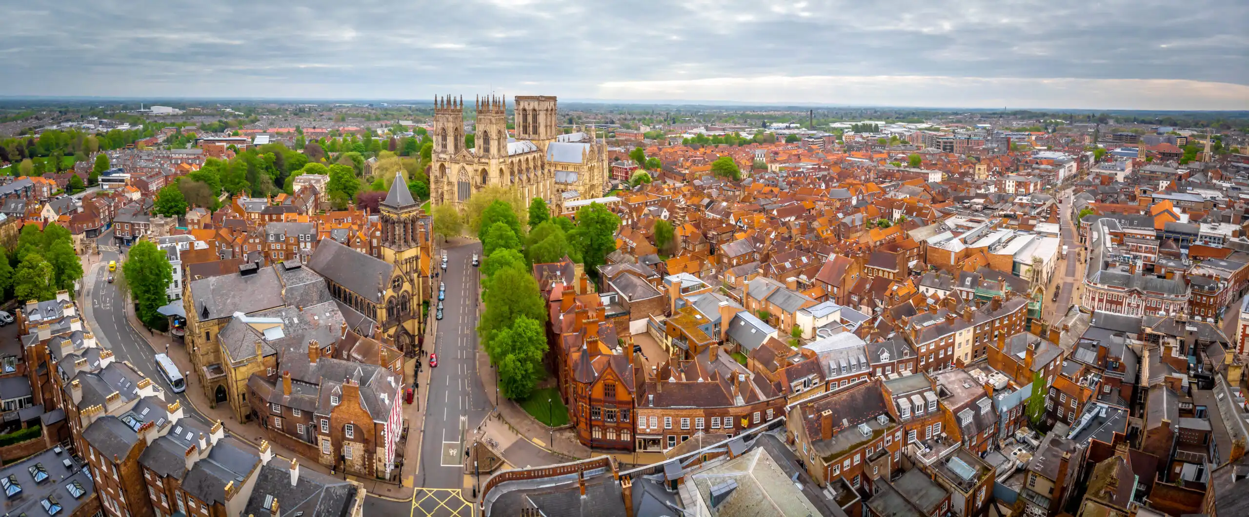 Aerial panoramic view of a historic town centered on a large gothic cathedral with red-tiled rooftops and tree-lined streets underneath a cloudy sky.