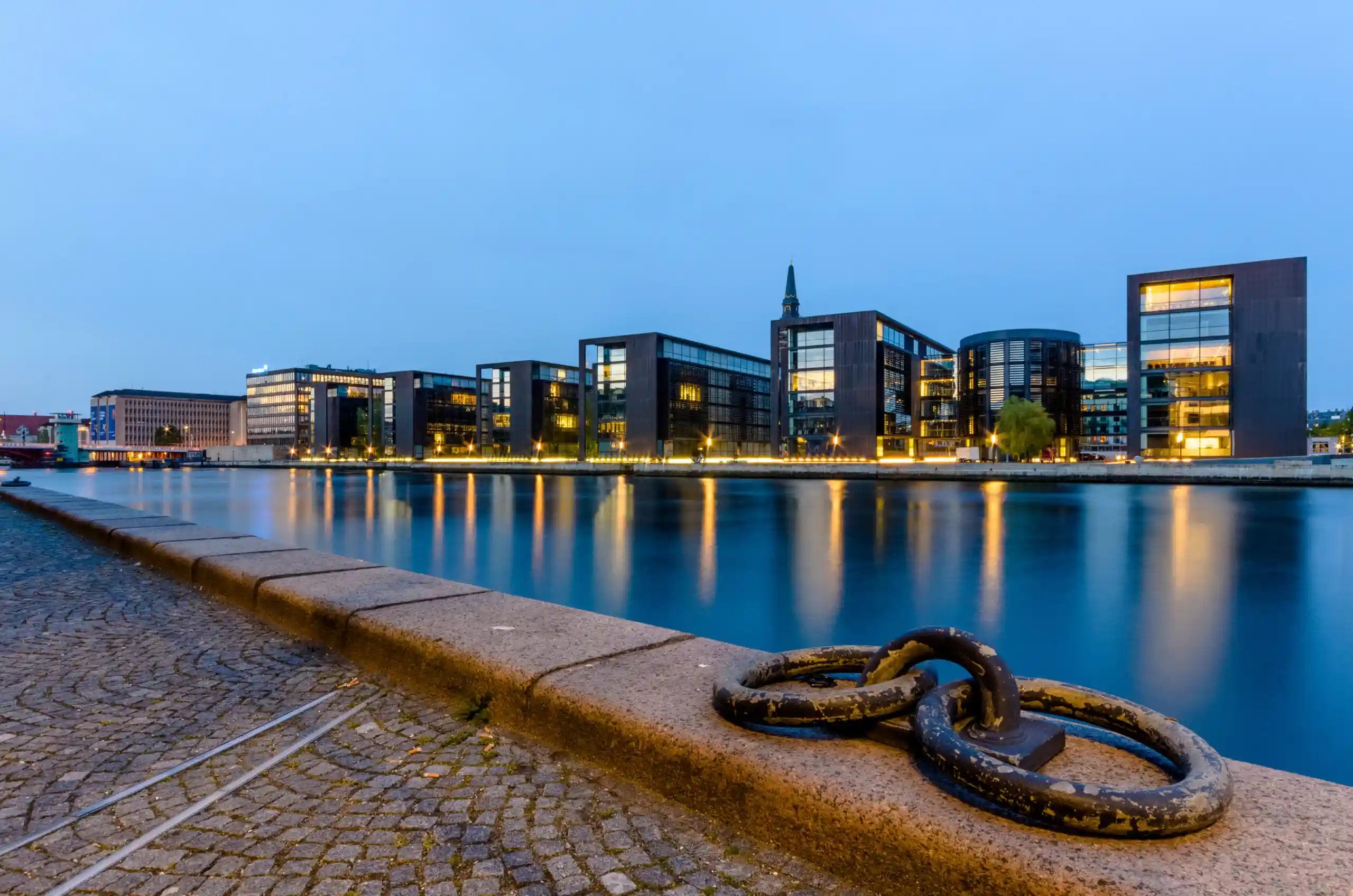 Evening view of modern glass buildings along a calm river with a cobblestone quay in the foreground and reflections in the water.